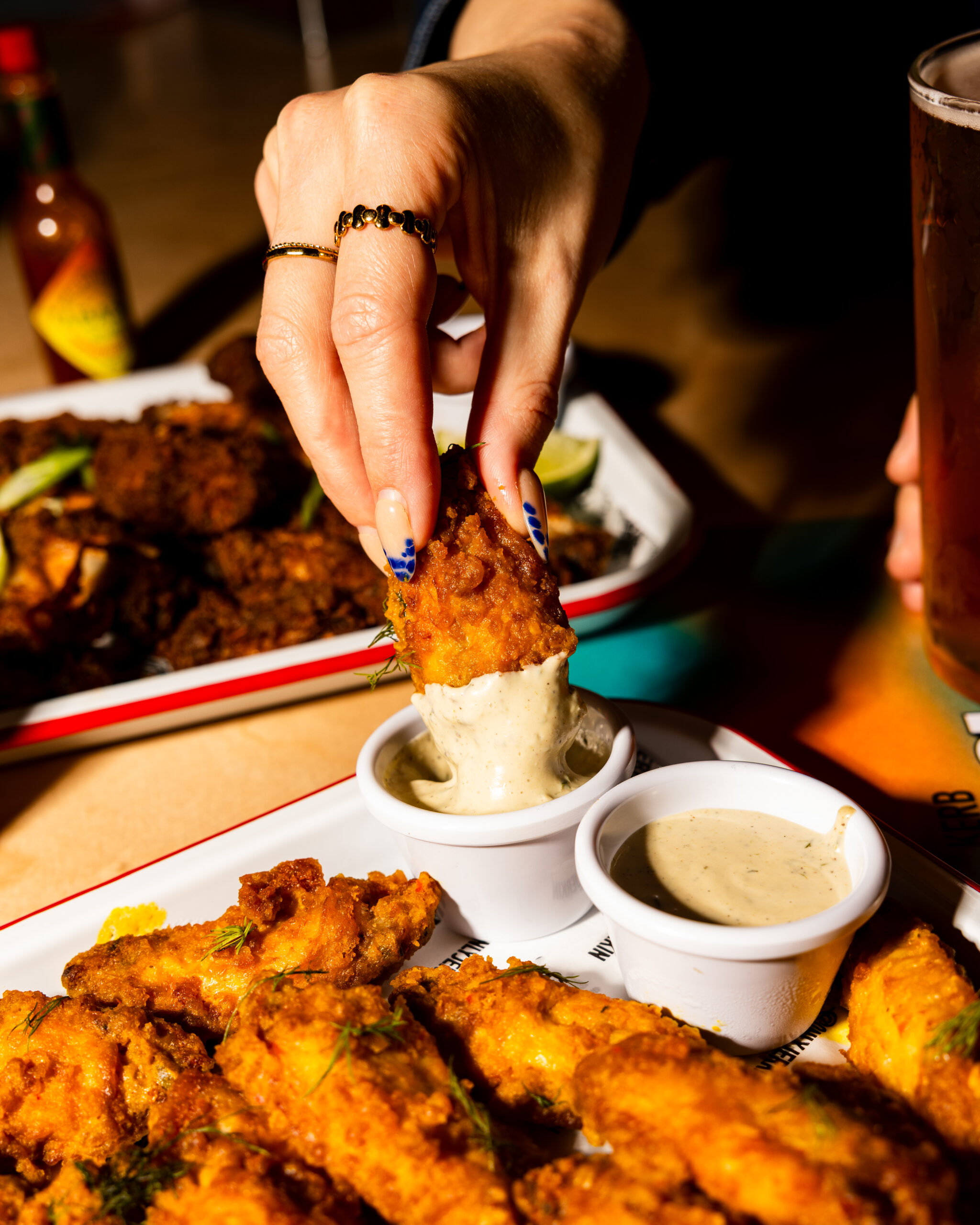 A hand with painted nails dips a crispy chicken wing into a cup of creamy dipping sauce, with more wings and a glass of beer on the table in a warmly lit setting.