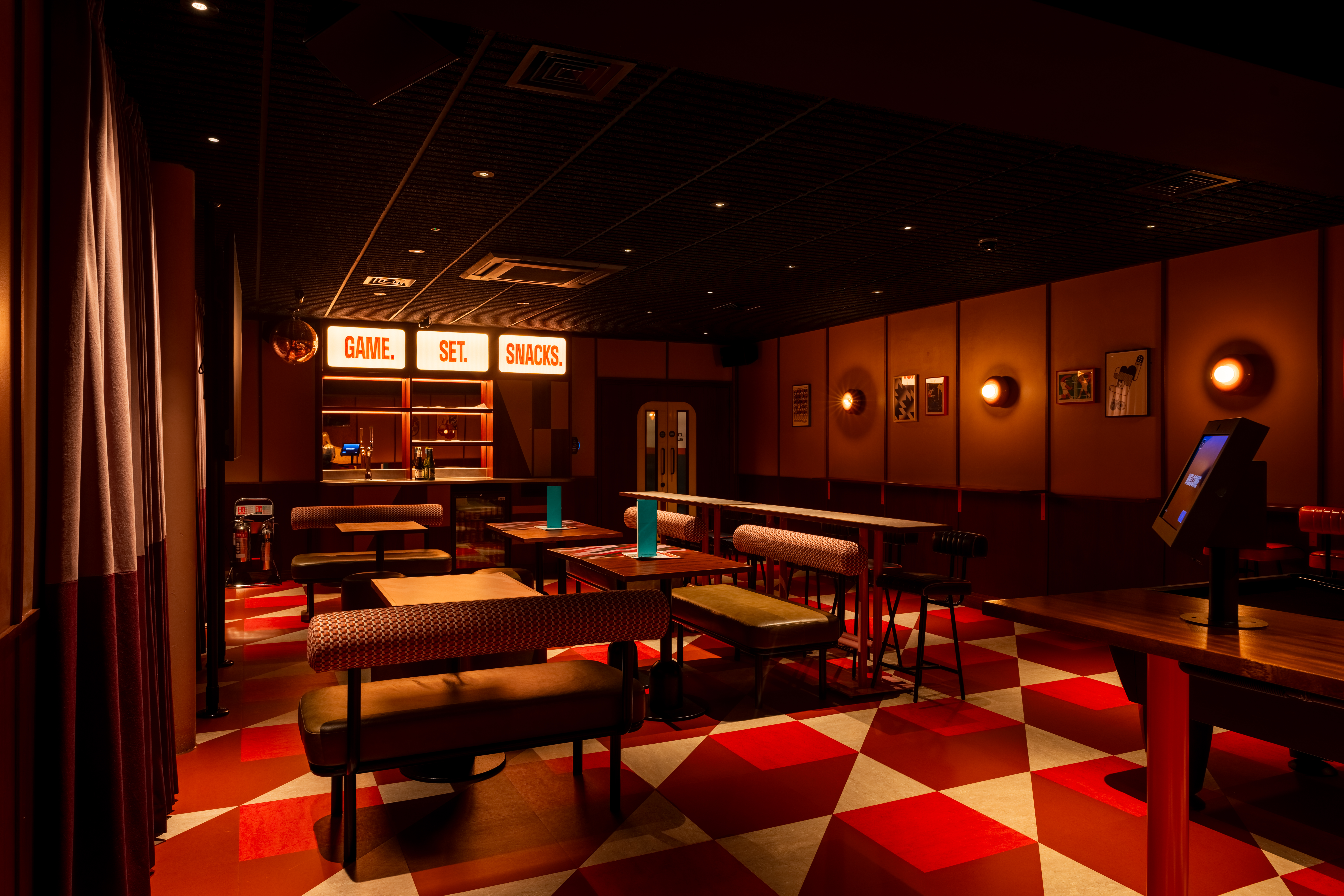 A cosy, dimly lit lounge with red and white chequered floors, modern tables and benches, and a bar area featuring illuminated Game. Set. Snacks. signs—perfect for catching every World Cup 2026 match in a relaxed, warm atmosphere.