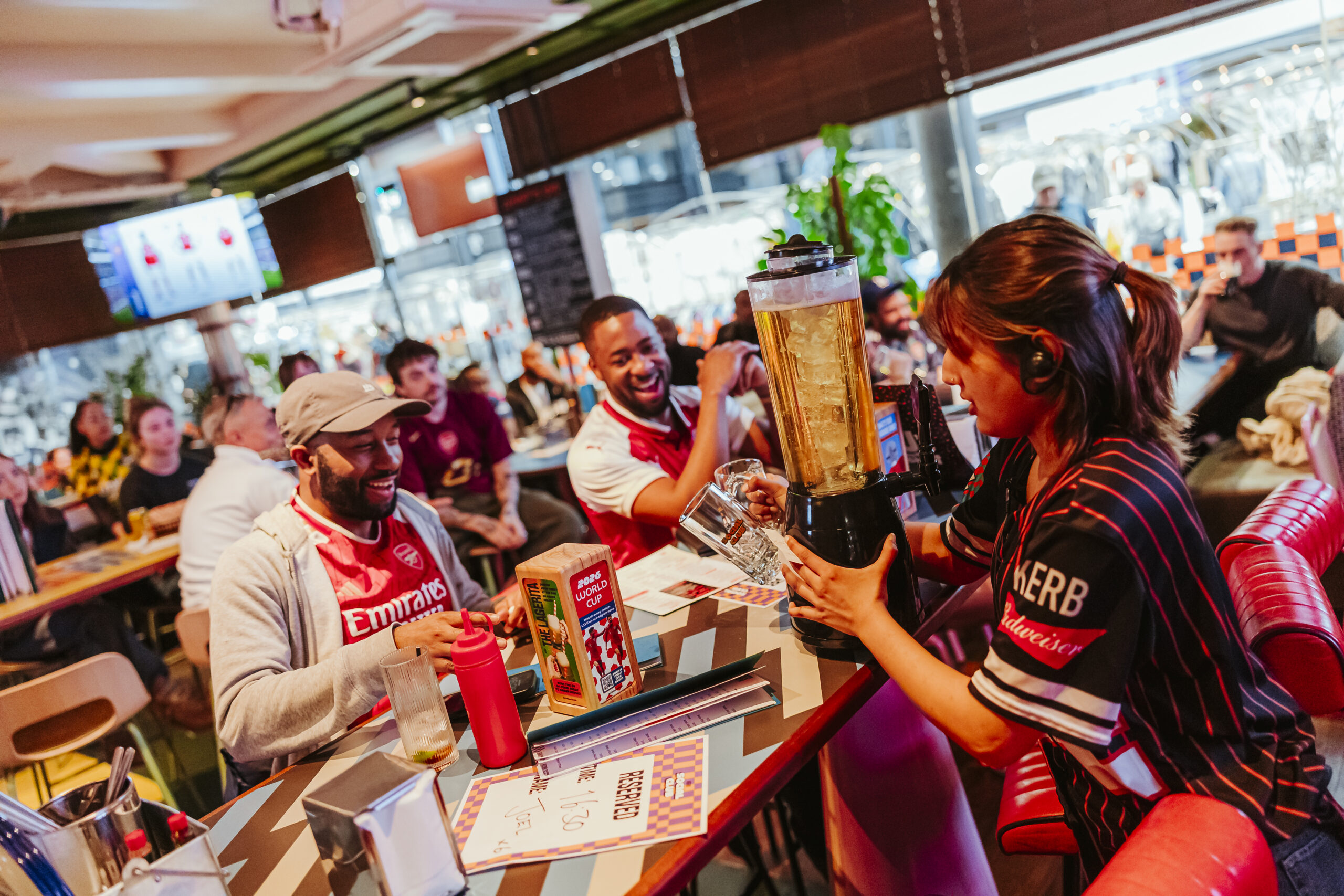 A woman serves up a tower of beer at a lively restaurant table, where three people in sports shirts are smiling and enjoying themselves, celebrating the excitement of World Cup 2026.