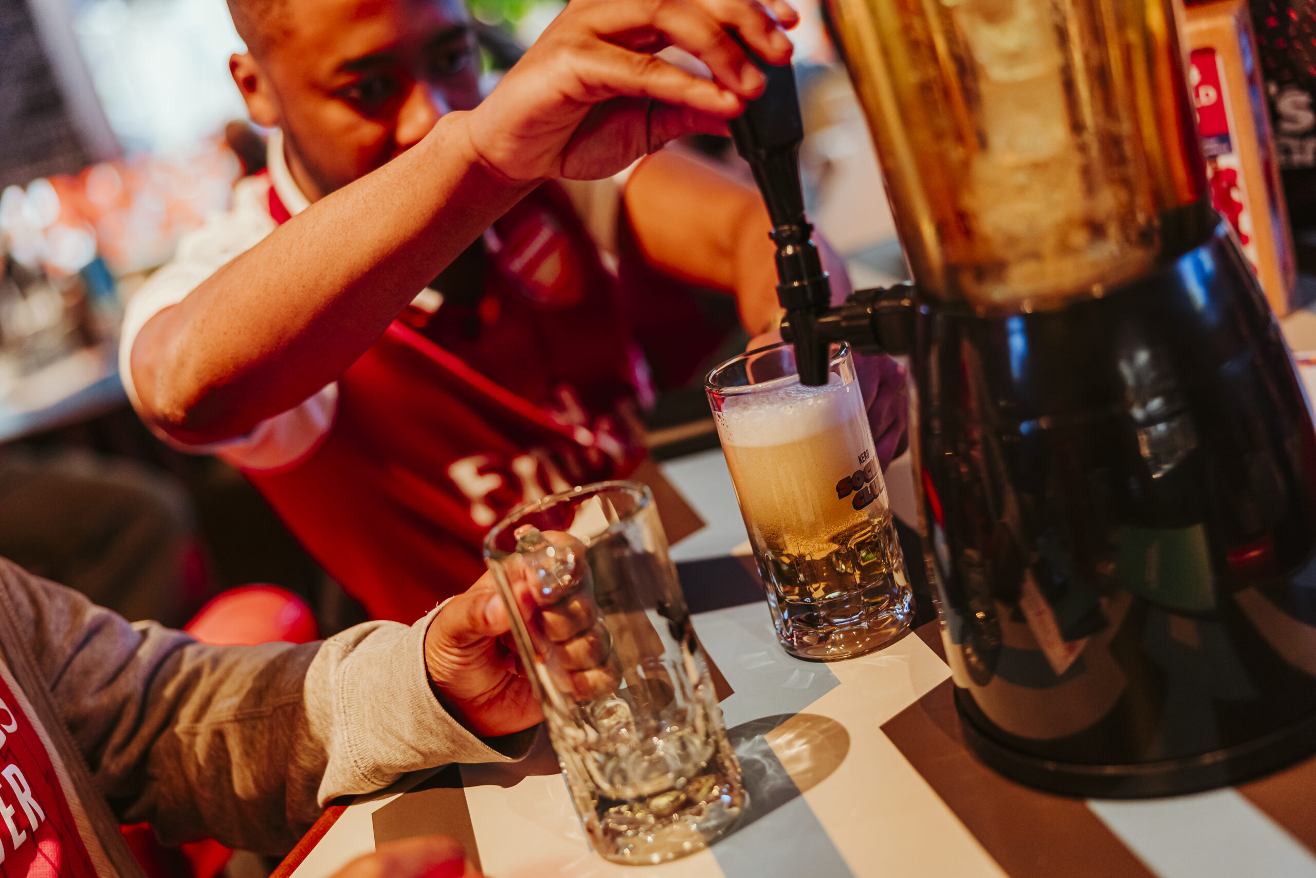 A person in a red sports shirt pours beer from a tabletop dispenser into a glass, with another empty glass nearby, as excitement for the World Cup 2026 fills the lively indoor setting.