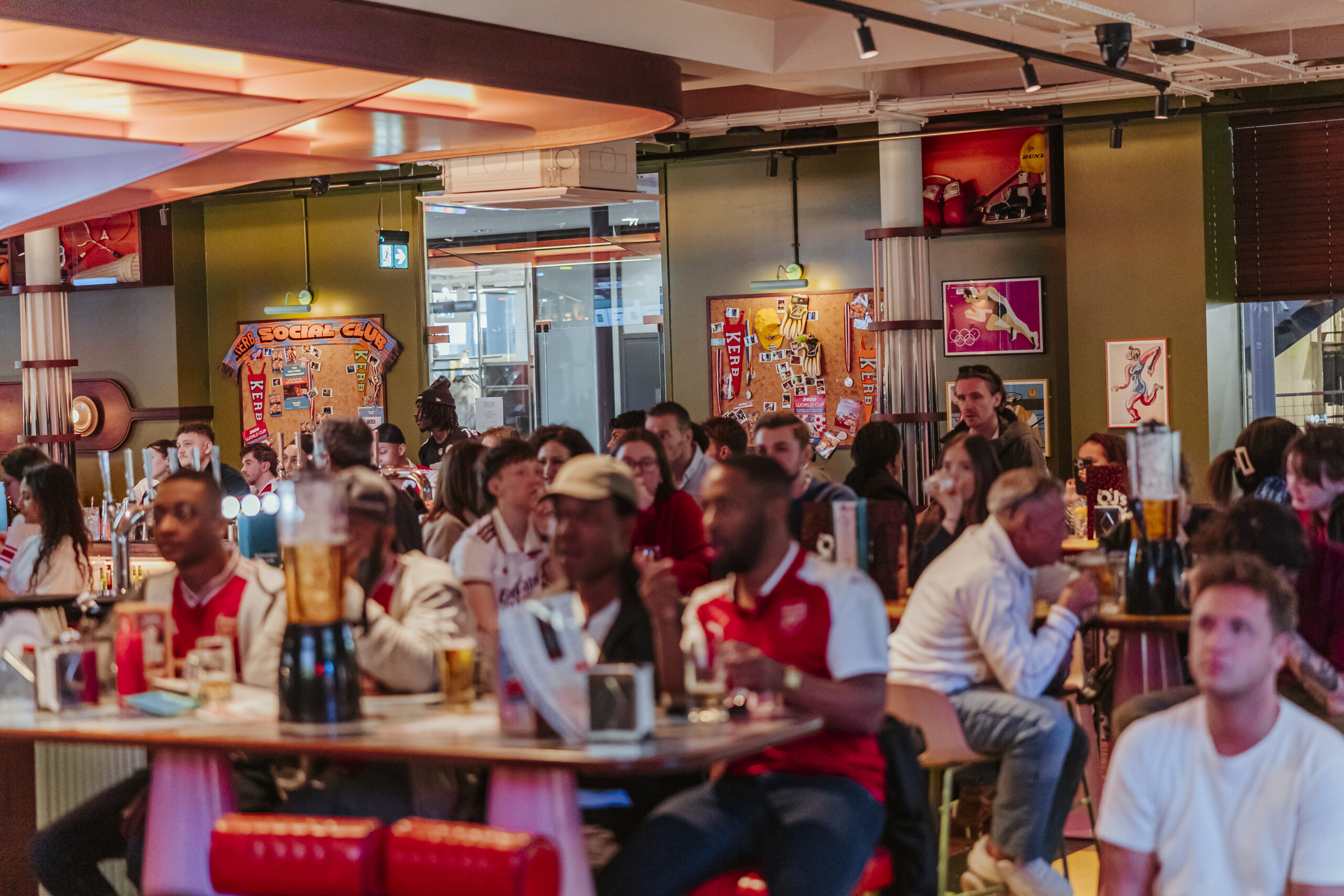A lively crowd of people, many in red and white sports jerseys, watch a World Cup 2026 event on screens in a bar decorated with football memorabilia. Drinks and snacks are on the tables, fuelling the energetic, engaged atmosphere.
