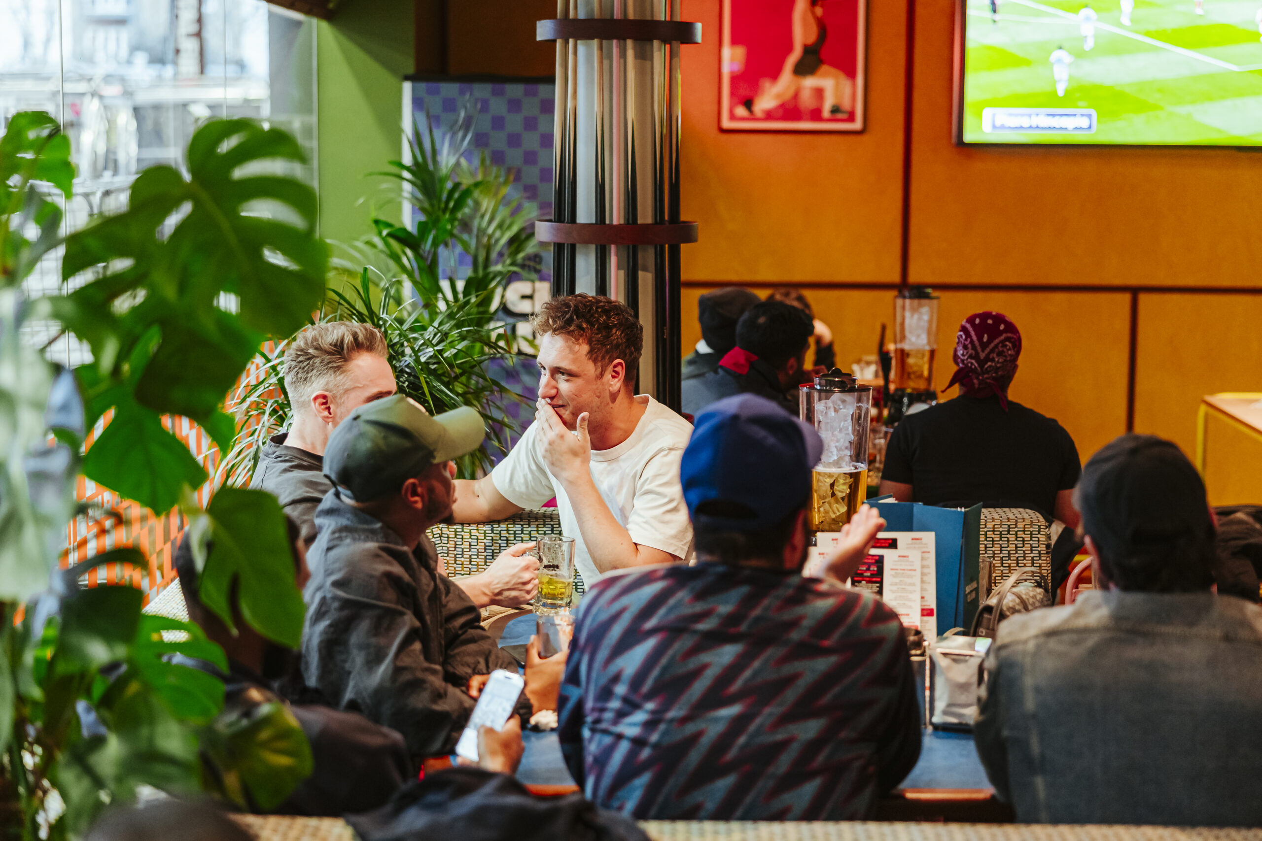 A group of people sit around a table in a lively bar or cafe, chatting and drinking as the World Cup 2026 match plays on a TV screen in the background. Green plants decorate the space.
