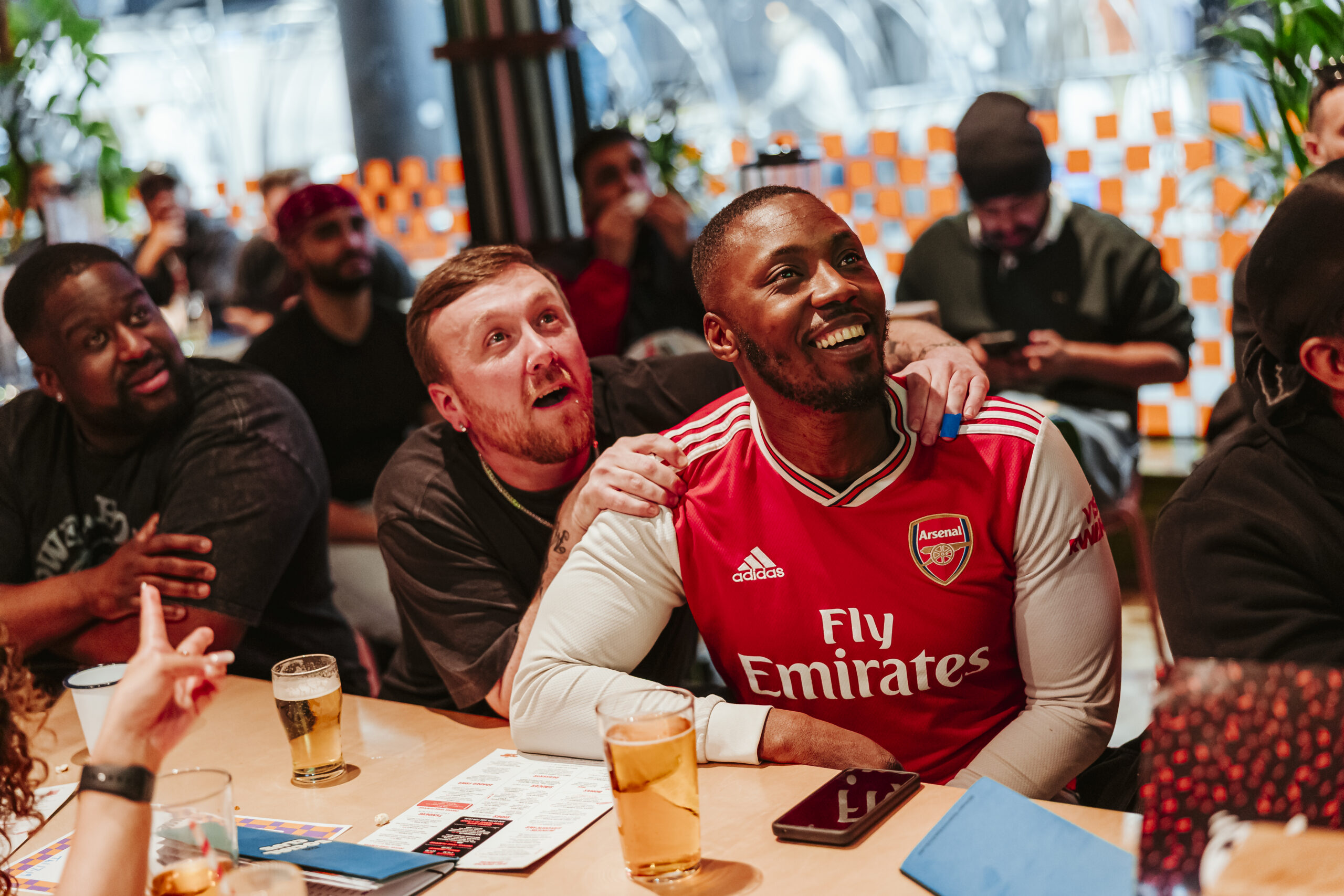 A group of people sit at a table, smiling and watching something off-camera. One man in an Arsenal shirt is at the centre, with another man’s arm around his shoulder. Drinks and menus are on the table as they chat about World Cup 2026 excitement.