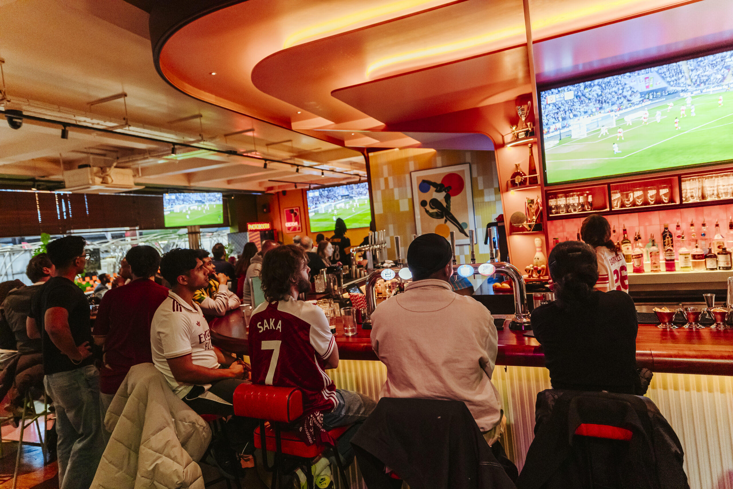 People sit at a busy bar, watching a World Cup 2026 football match on large TVs. Drinks line the counter and fans in kits, including one with Saka 7, create a lively atmosphere focused on the excitement of the game.